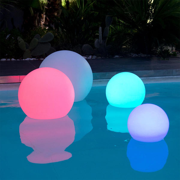 Vier leuchtende Kugeln in Pink, Weiß und Blau schwimmen nachts in einem Pool mit Garten im Hintergrund.
