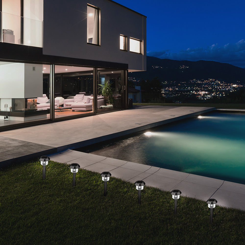 Sechs solarbetriebene Wegeleuchten leuchten im Gras neben einem modernen Pool vor einem beleuchteten Haus bei Nacht.