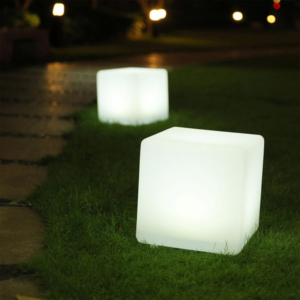Zwei leuchtende, weiße würfelförmige Lampen stehen nachts auf grünem Gras neben einem gepflasterten Weg.