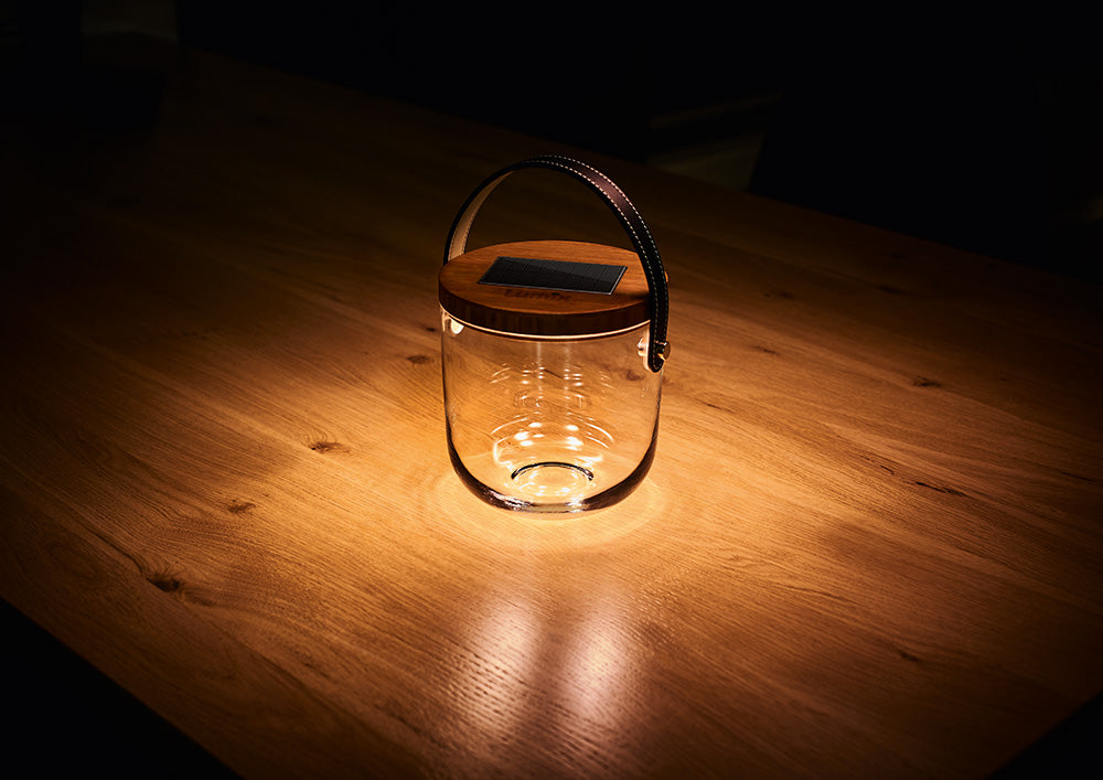 Eine elegante Glaslaterne mit Holzigel und Ledergriff steht auf einem warm beleuchteten Holztisch.