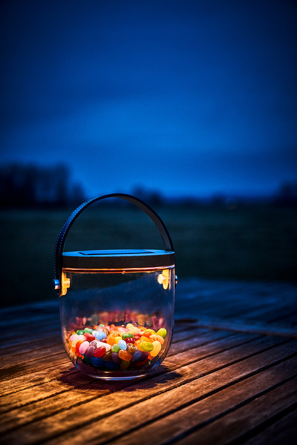 Beleuchtetes Glasgefäß mit bunten Geleebonbons auf einem Holztisch bei Dämmerung.