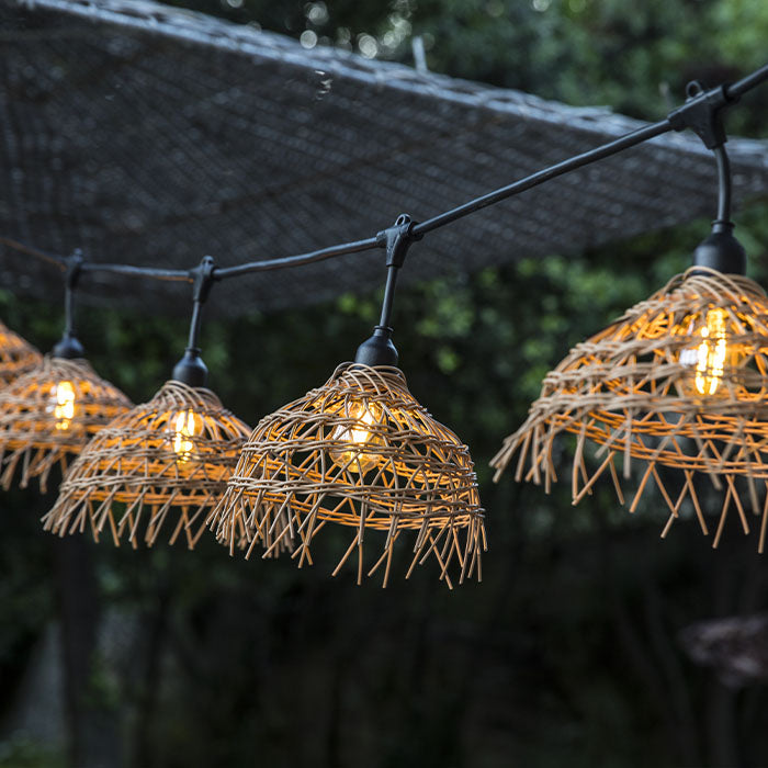 Eine Lichterkette mit geflochtenen Rattanschirmen leuchtet sanft vor einem dunklen Hintergrund.