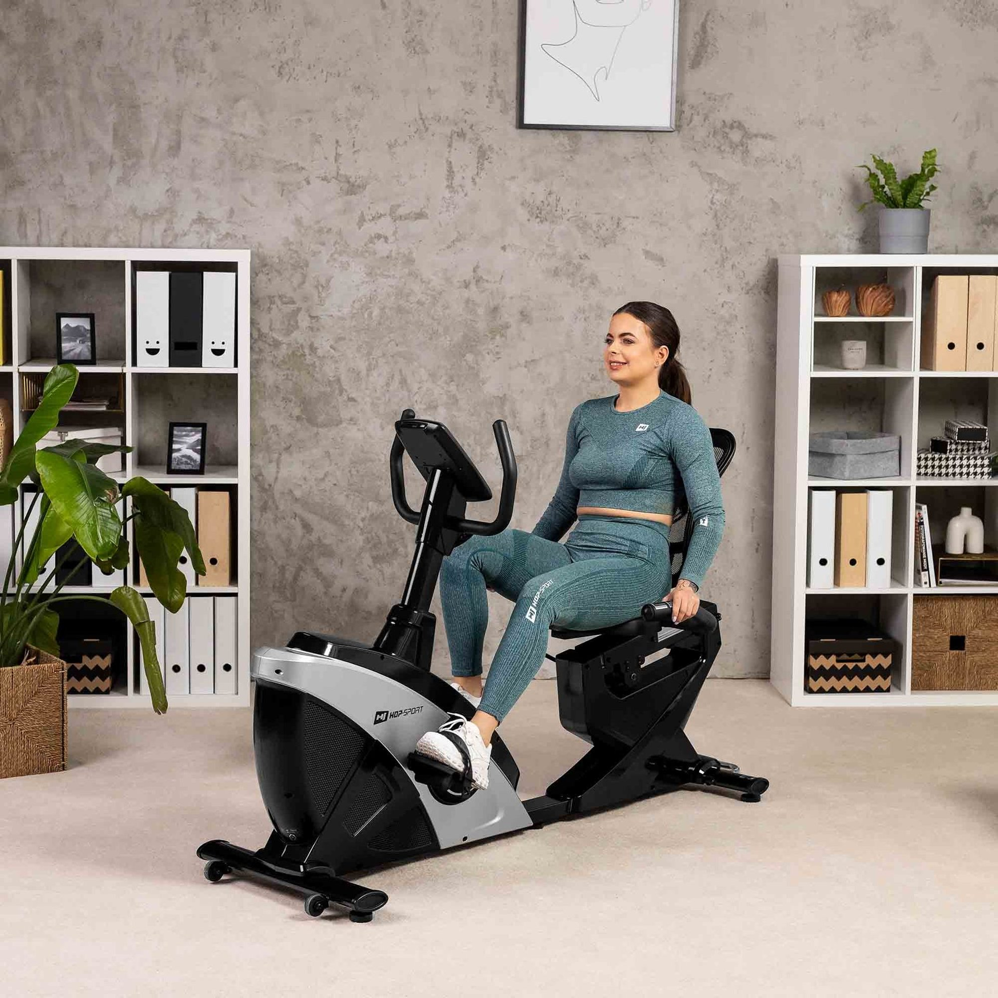 Eine Frau in blauer Sportkleidung trainiert auf einem schwarzen Liegeheimtrainer in einem modern eingerichteten Zimmer.