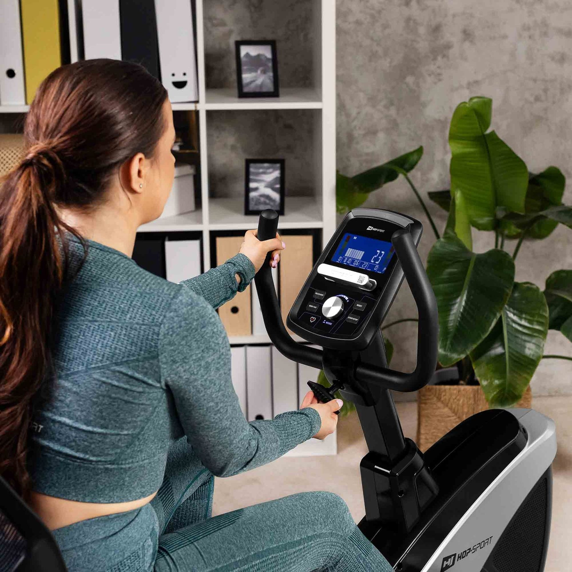 Eine Frau in Sportkleidung trainiert auf einem Heimtrainer vor einem Regal mit Pflanzen.