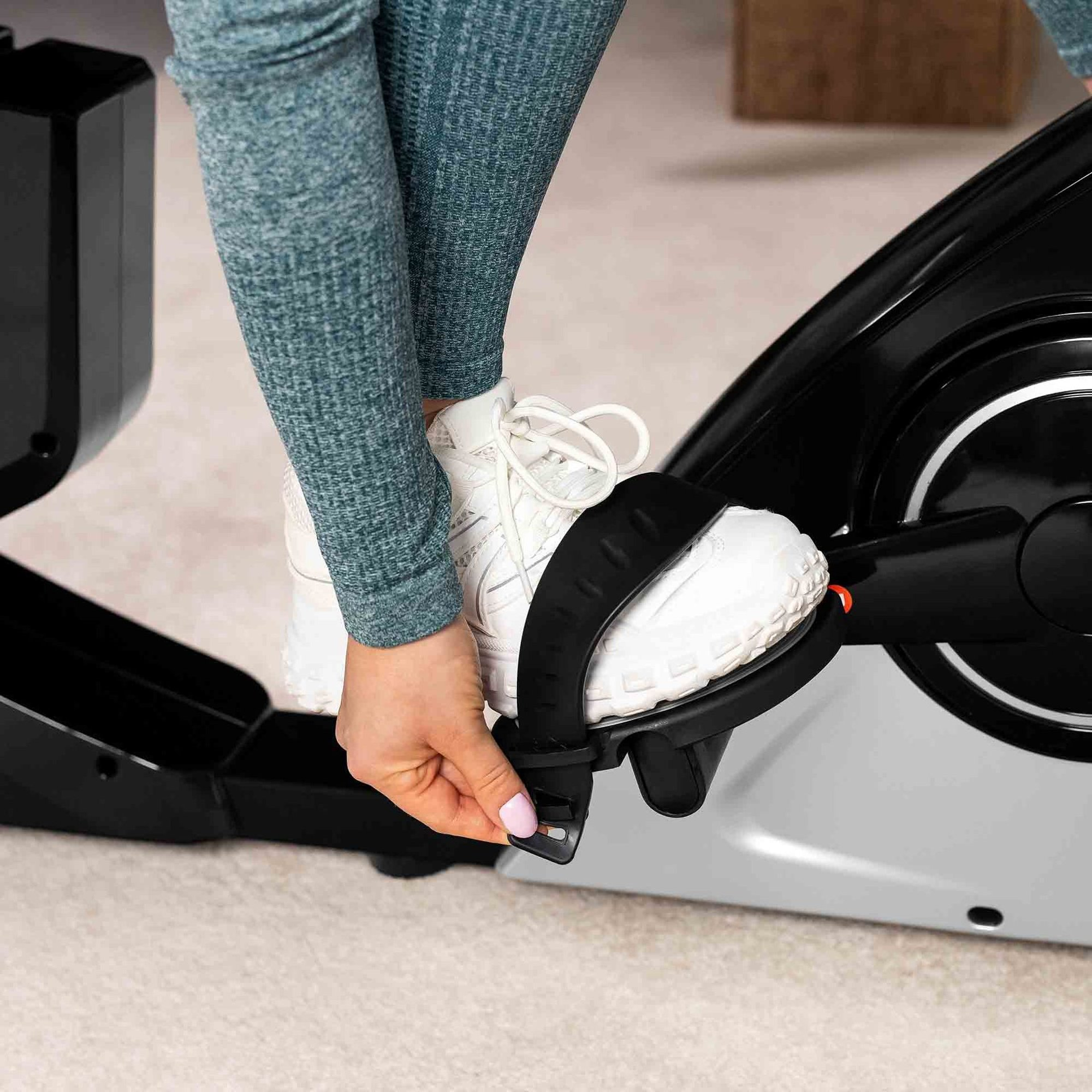 Person in blauer Legging und weißen Sneakers adjustiert den Riemen eines schwarzen Heimtrainerpedals.