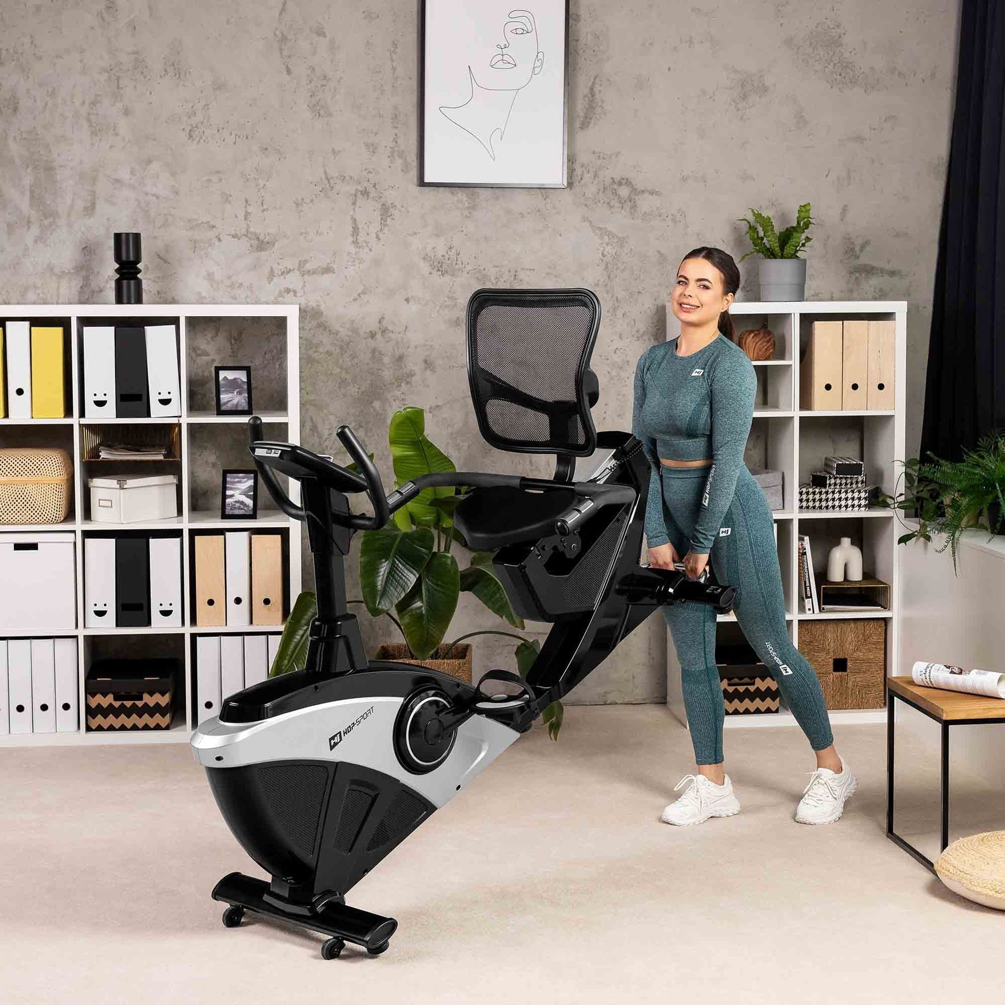 Eine lächelnde Frau in türkisfarbener Sportkleidung steht neben einem Liegeheimtrainer in einem Wohnzimmer.