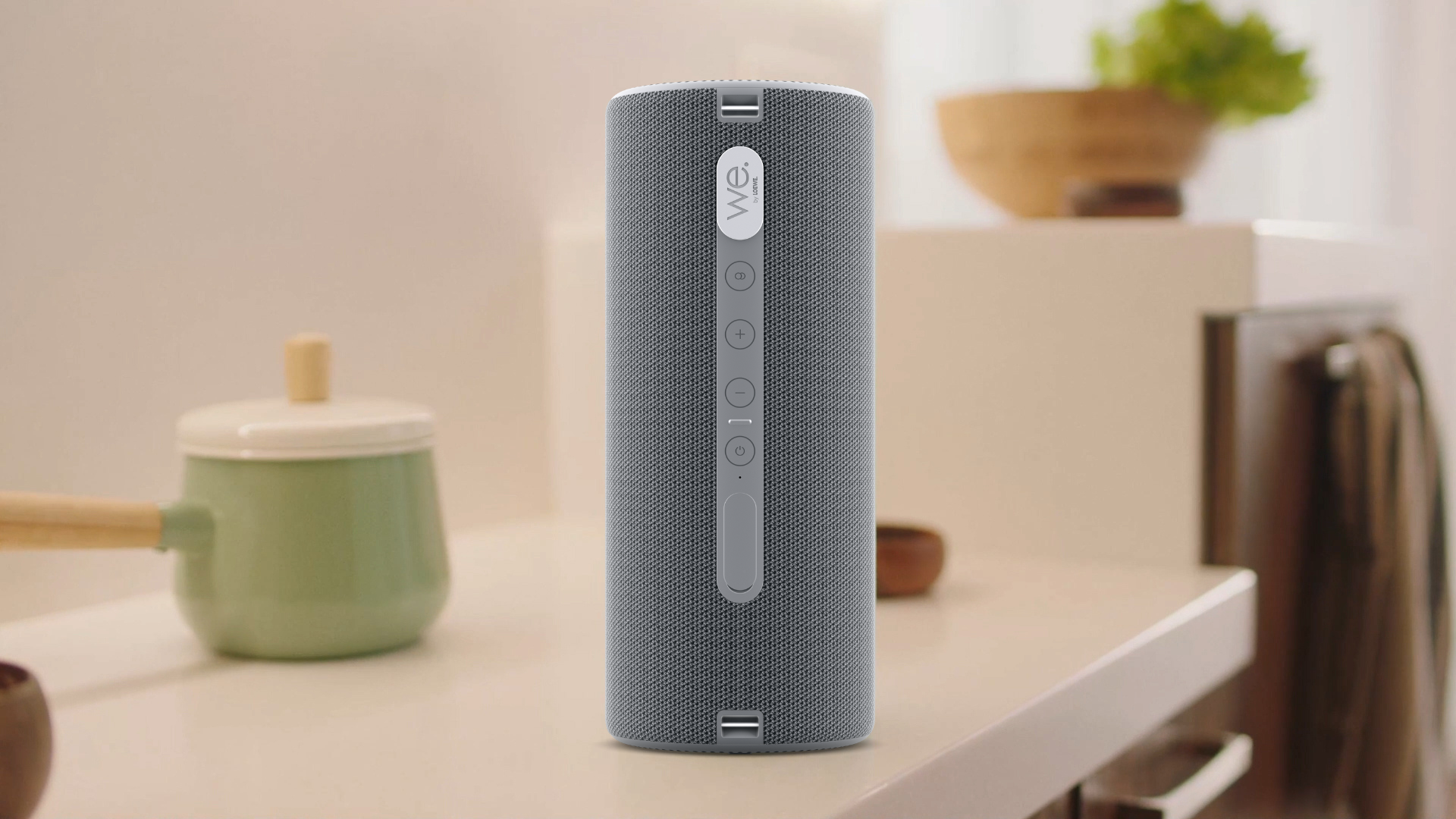 Ein grauer zylindrischer Bluetooth-Lautsprecher steht auf einer hellen Küchentheke vor einem verschwommenen Hintergrund.
