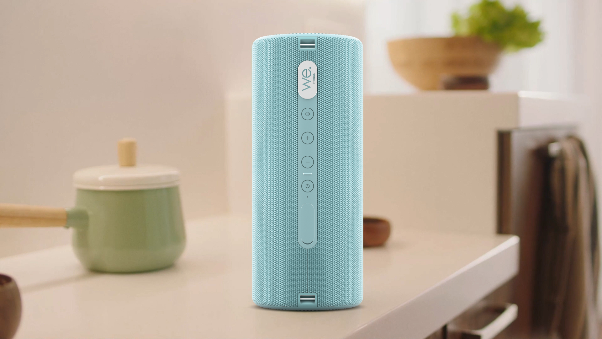 Ein hellblauer zylindrischer Bluetooth-Lautsprecher steht auf einer weißen Theke in einer Küche.