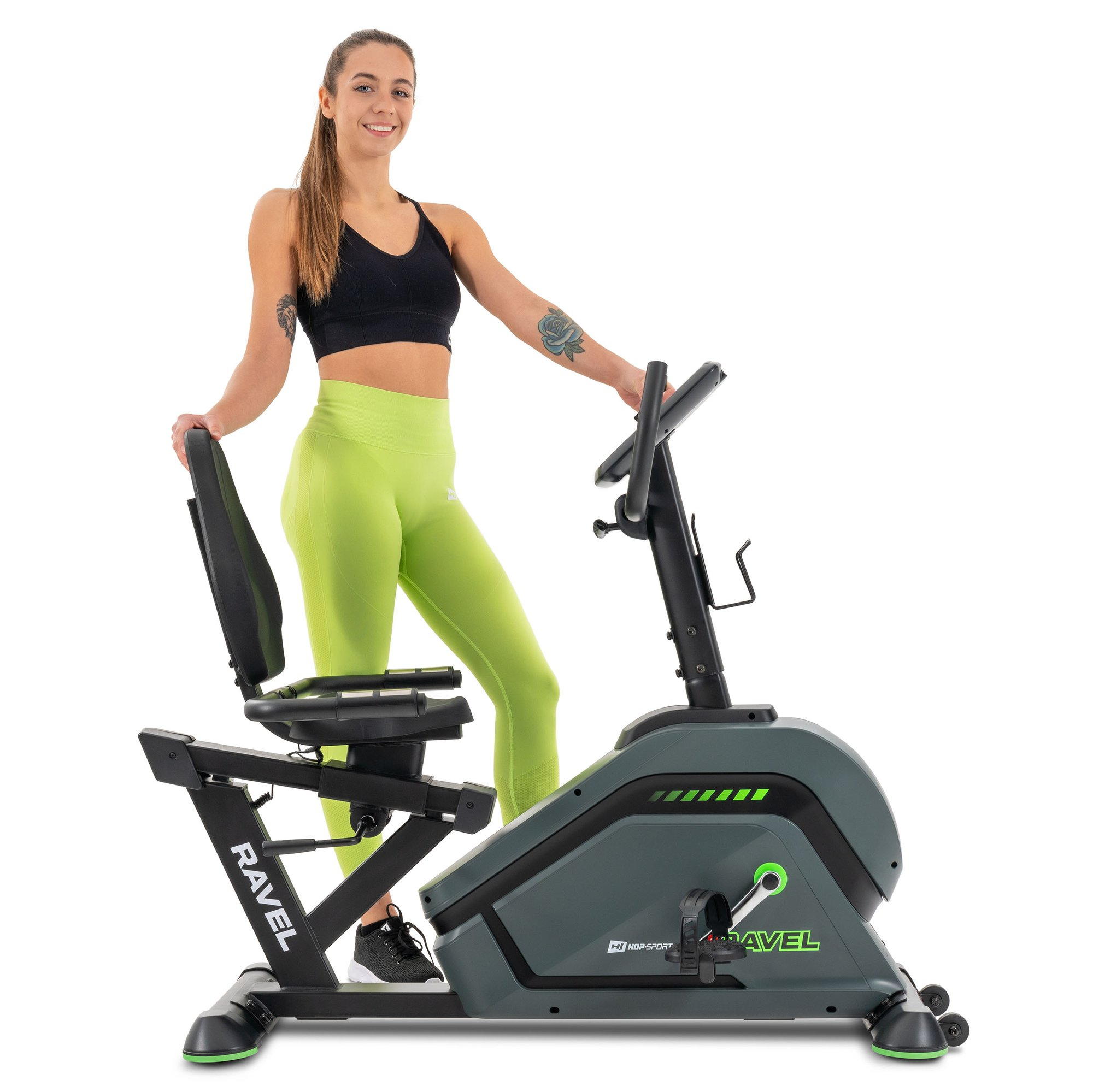 Eine lächelnde Frau in Sportkleidung steht vor weißem Hintergrund neben einem schwarz-grünen Liegeheimtrainer.
