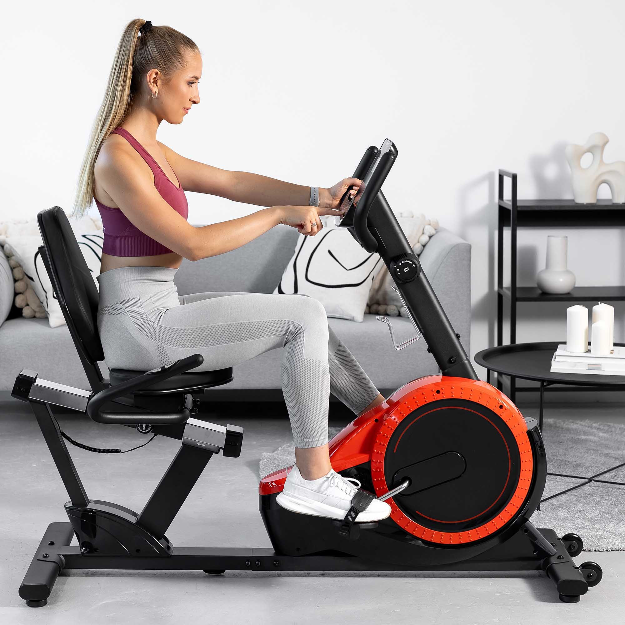 Eine blondhaarige Frau in Sportkleidung trainiert auf einem roten Heimtrainer in einem modernen Wohnzimmer.