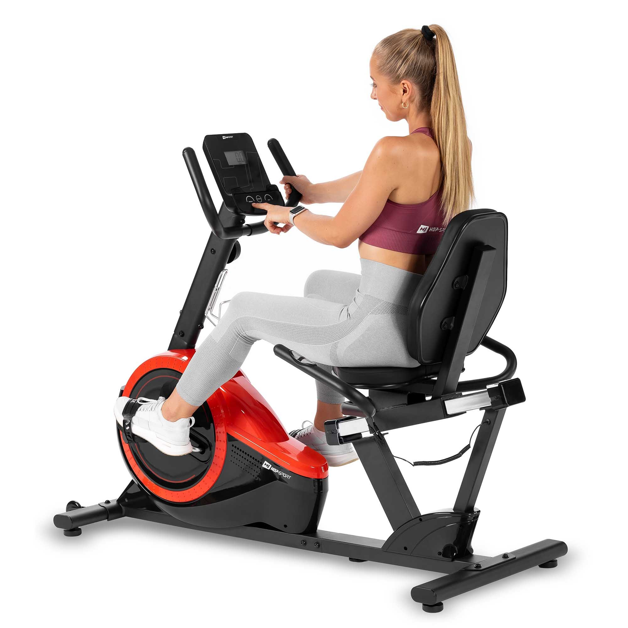 Eine blonde Frau sitzt auf einem roten und schwarzen Liegeheimtrainer und bedient das Display.