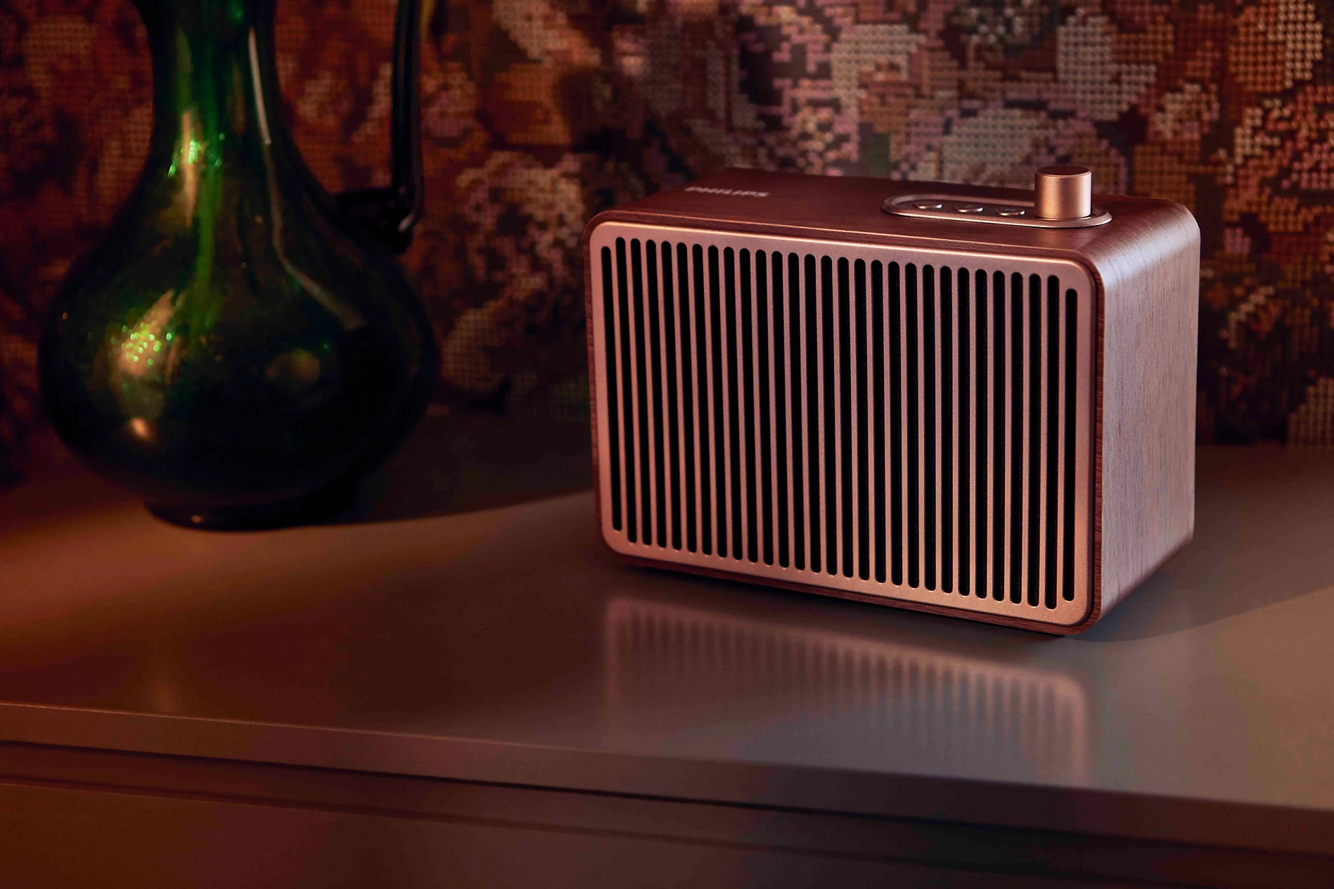 Dikey ızgaralı retro kahverengi Philips hoparlör, yeşil cam vazonun yanında masada duruyor.