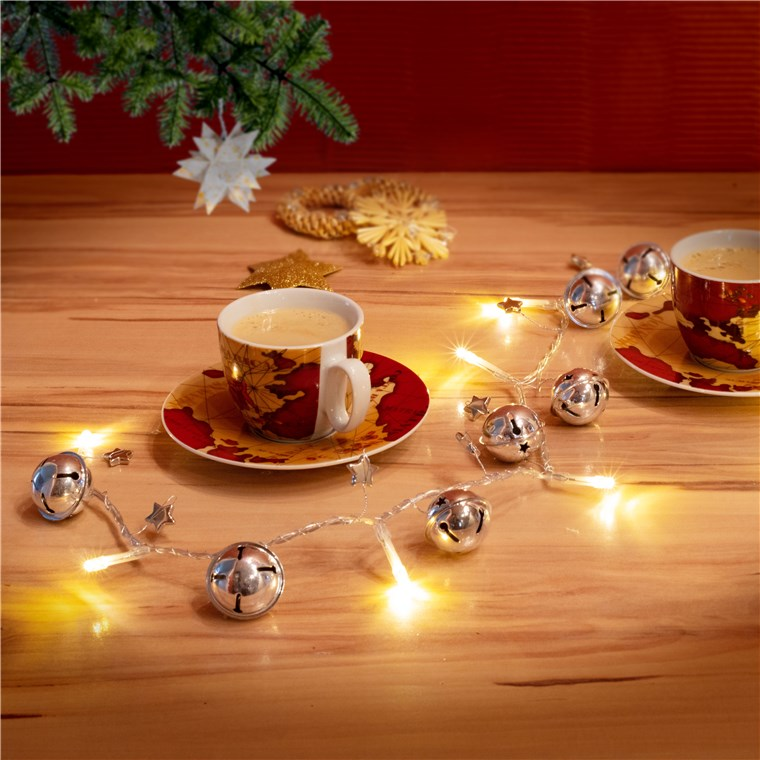 Kaffeetassen, eine Lichterkette mit Glocken und Weihnachtsdekoration auf einem Holztisch.