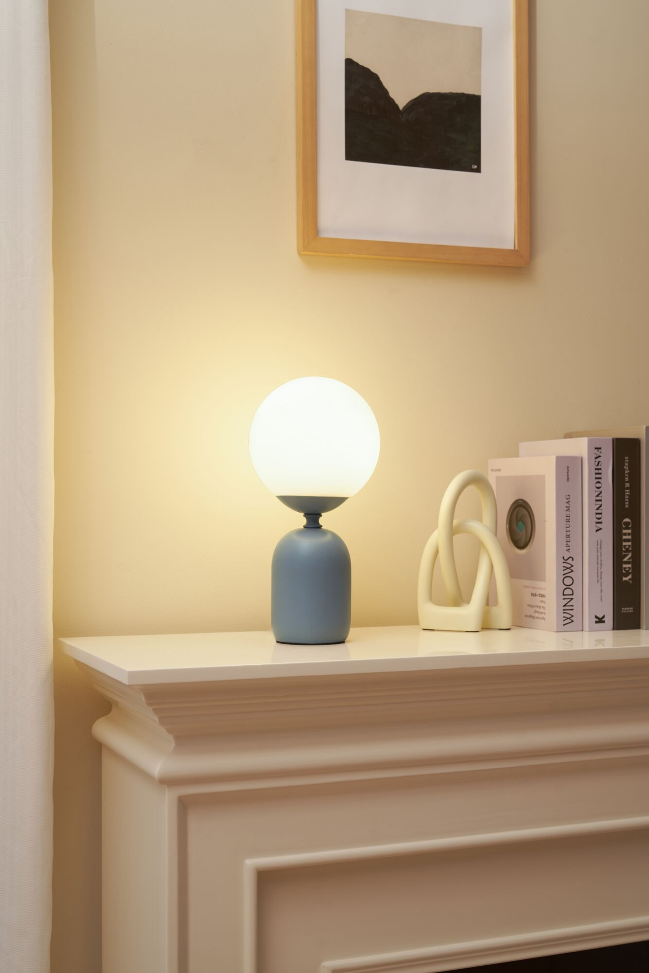 Eine blaue Kugel Tischlampe auf einem weißen Kaminsims neben Büchern und einer Skulptur.