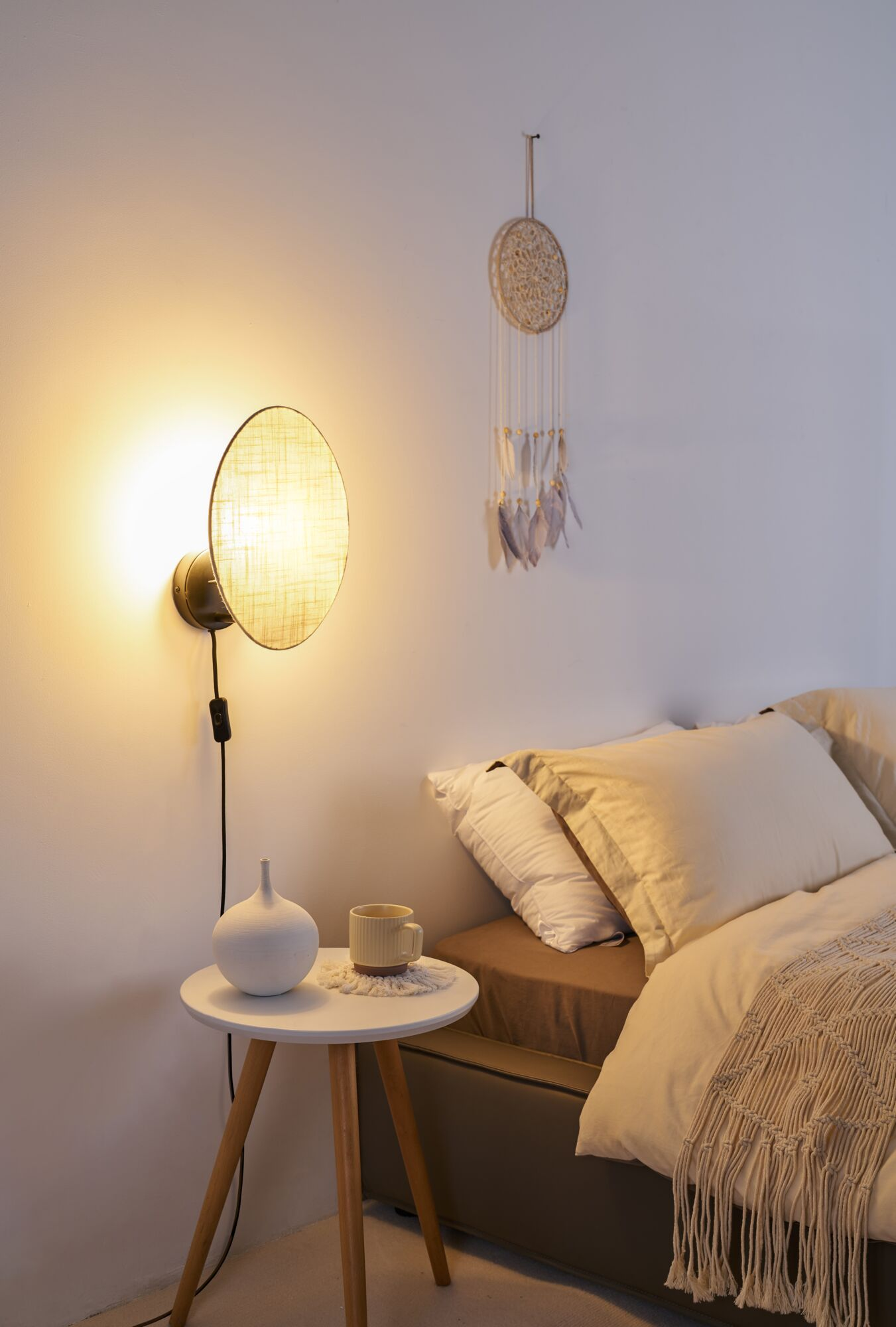 Ein weißer Nachttisch mit Lampe und Vase steht neben einem gemütlichen Bett, darüber hängt ein Traumfänger.