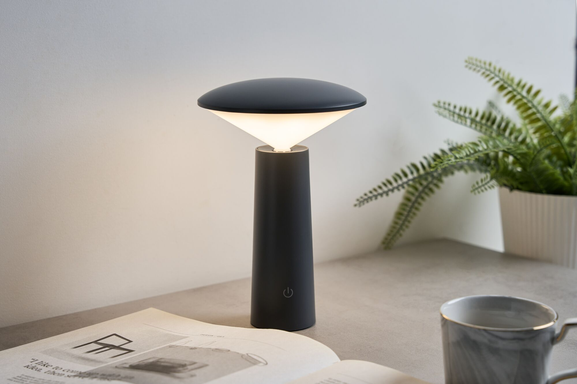 Moderne schwarze Tischlampe mit warmem Licht auf einem Tisch neben einer Zeitung und einer Kaffeetasse.