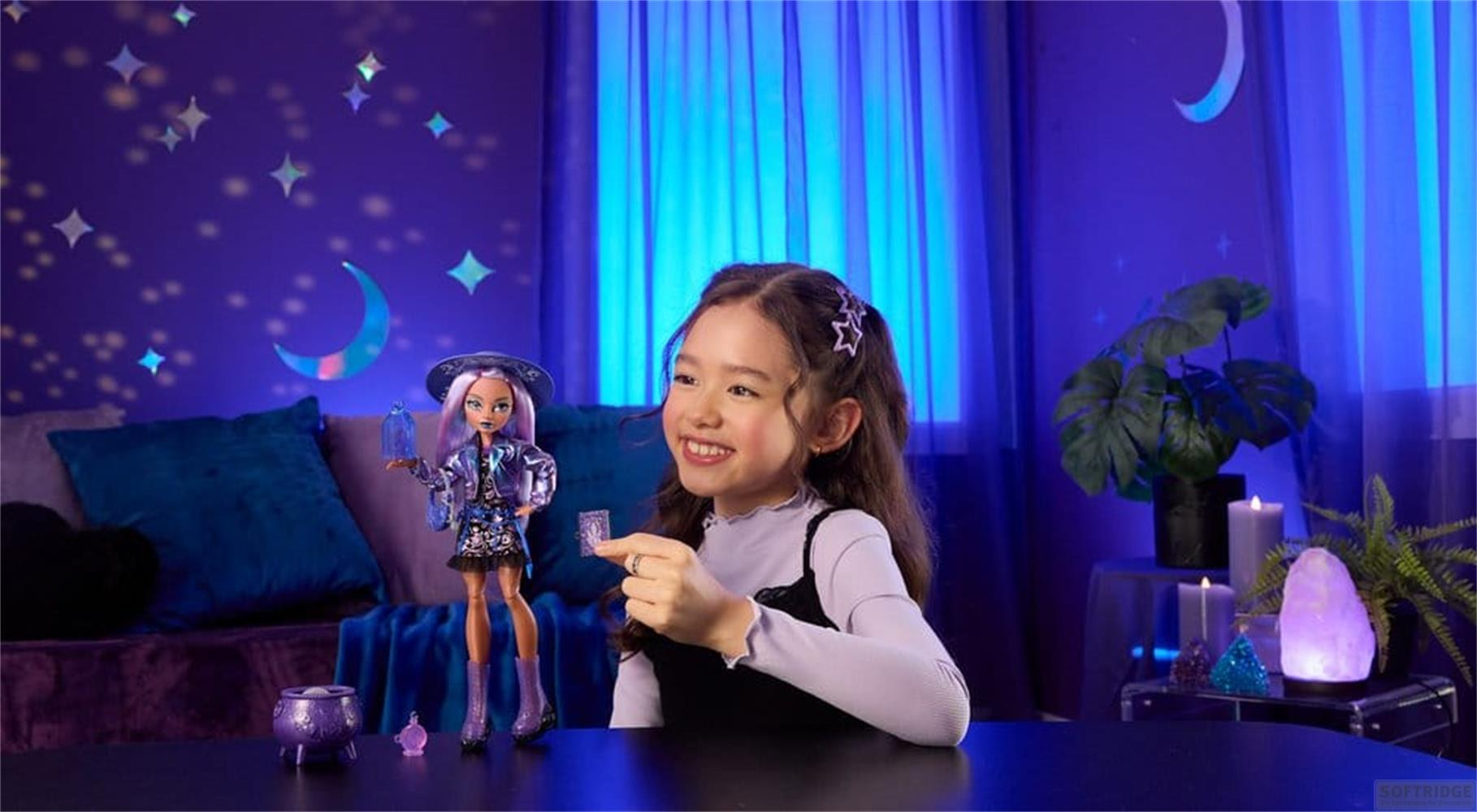 Una niña sonriente está sentada en una mesa con una muñeca, ante una pared azul con calcomanías de luna y estrellas.