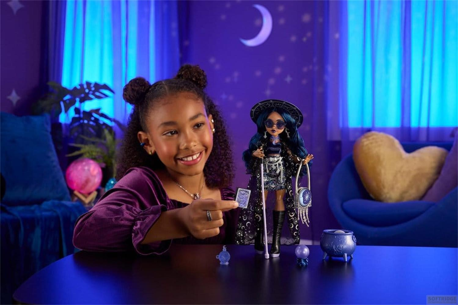 Una niña sonriente sostiene una pequeña tarjeta junto a una muñeca de cabello azul en una habitación iluminada de púrpura.
