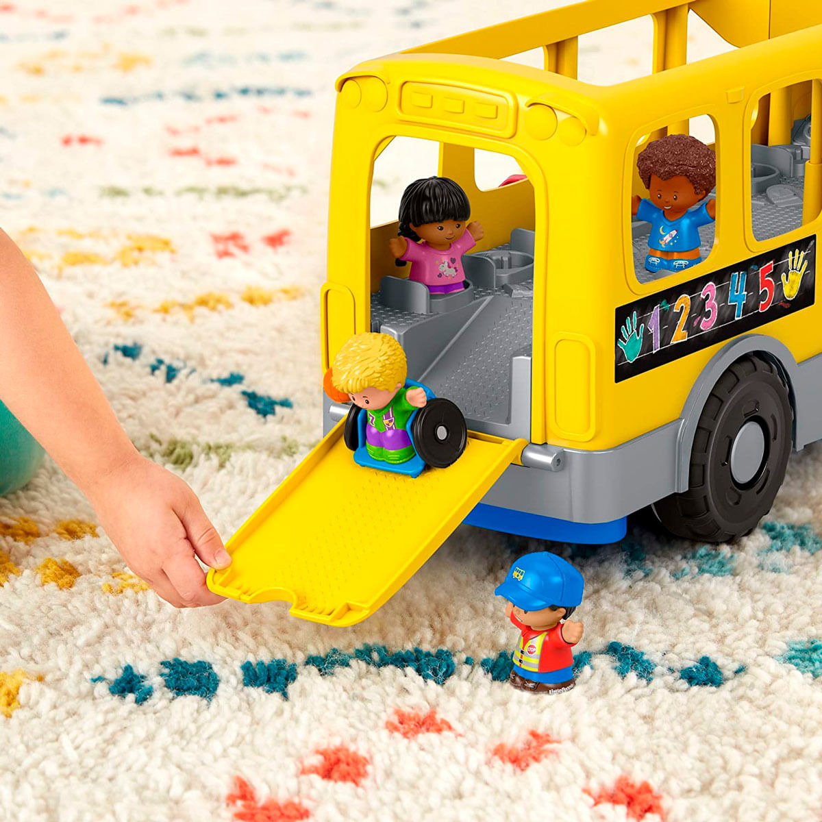 La mano de un niño baja una rampa de un autobús escolar de juguete amarillo con cuatro figuras en una alfombra colorida.