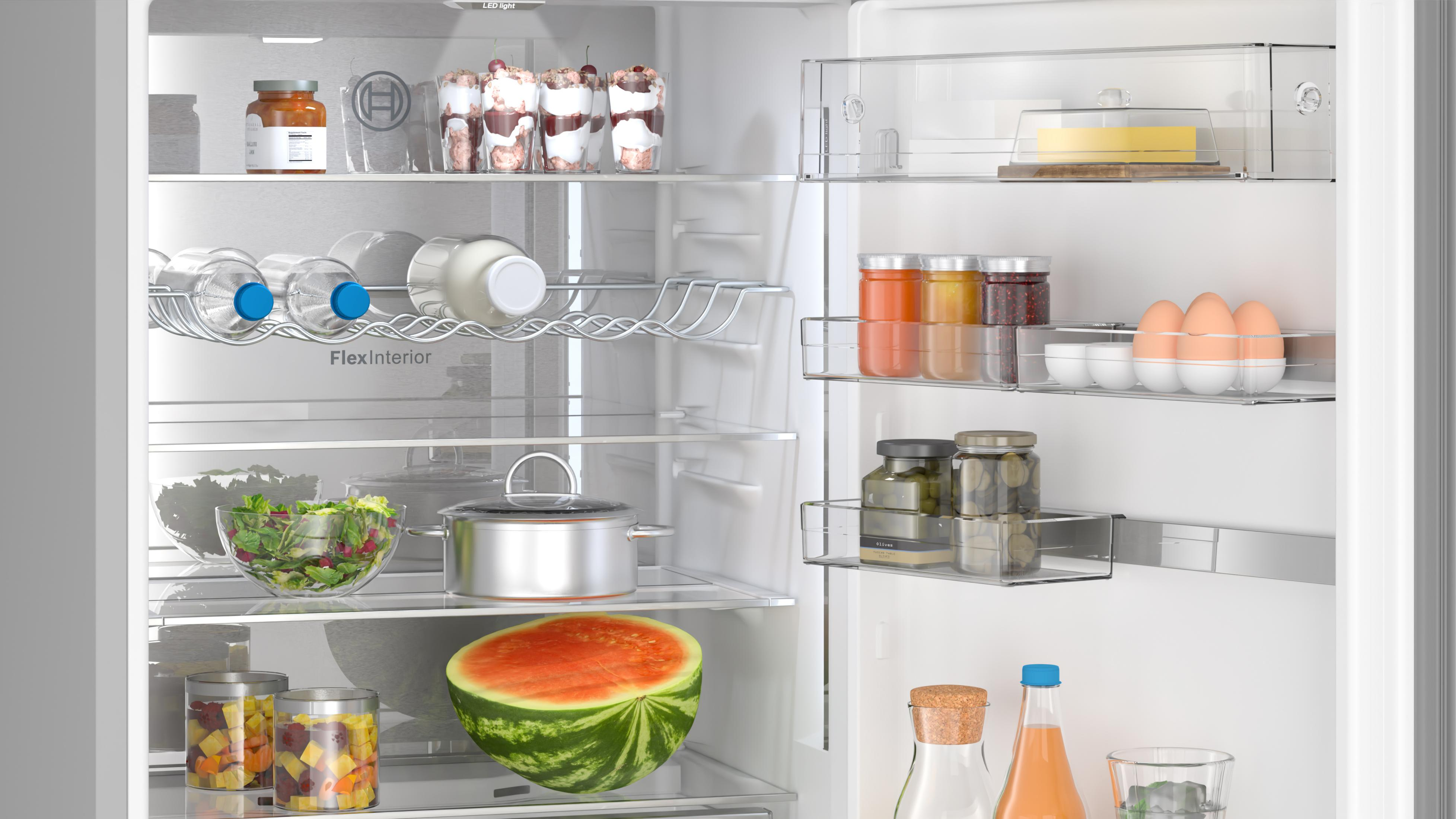Offener weißer Kühlschrank mit Lebensmitteln wie Salat, Topf, Obst, Flaschen und Eiern in Regalen.
