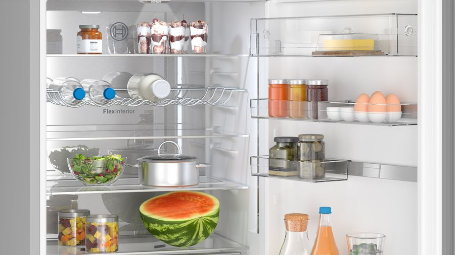 Geöffnete weiße Innenseite eines Kühlschranks, gefüllt mit Lebensmitteln, Gläsern, Flaschen, Salat und einer Wassermelone.