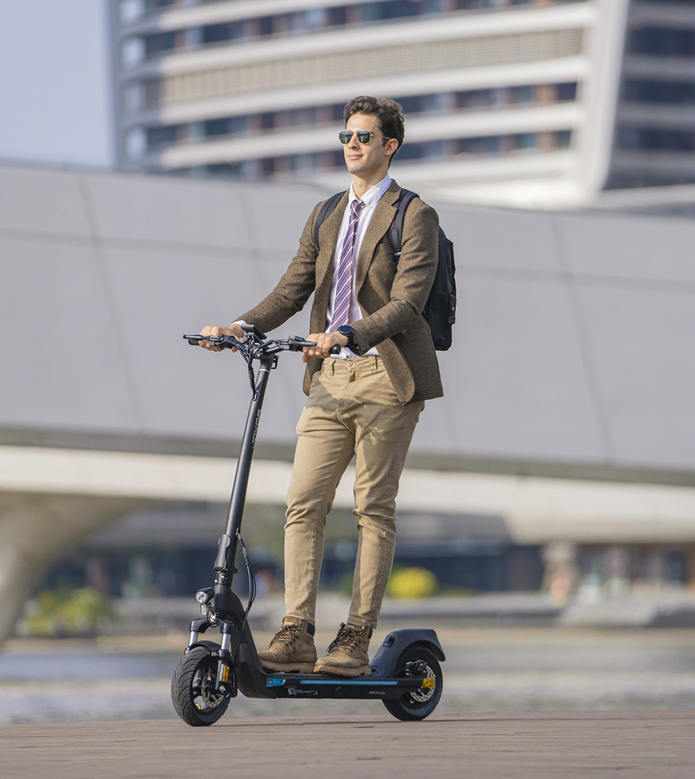 Un uomo in abito guida un monopattino elettrico davanti a edifici moderni.
