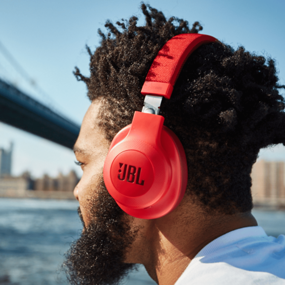 Un uomo barbuto con cuffie JBL rosse guarda l'acqua, con un ponte sullo sfondo.