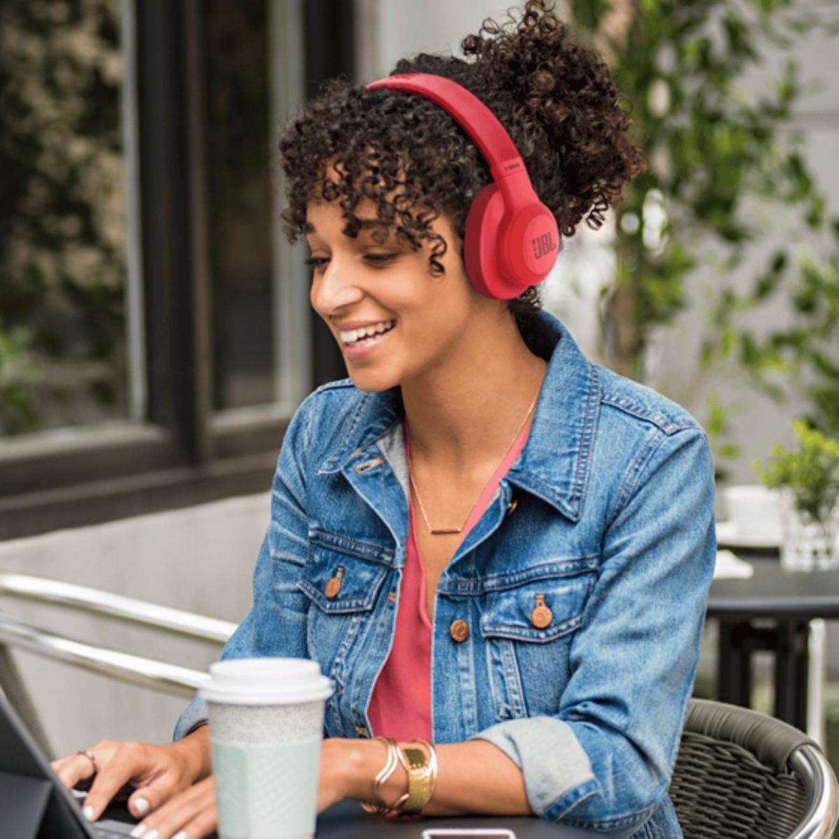 Una donna sorridente con cuffie rosse e giacca di jeans scrive su un tablet all'aperto.
