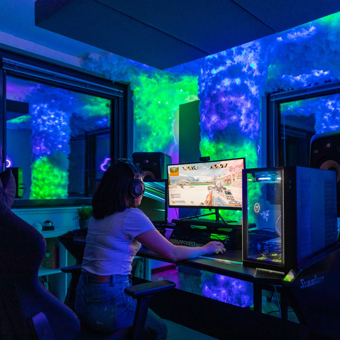 Eine Spielerin sitzt vor einem beleuchteten PC in einem Raum mit blauer Wolkenglut und grünem Licht.