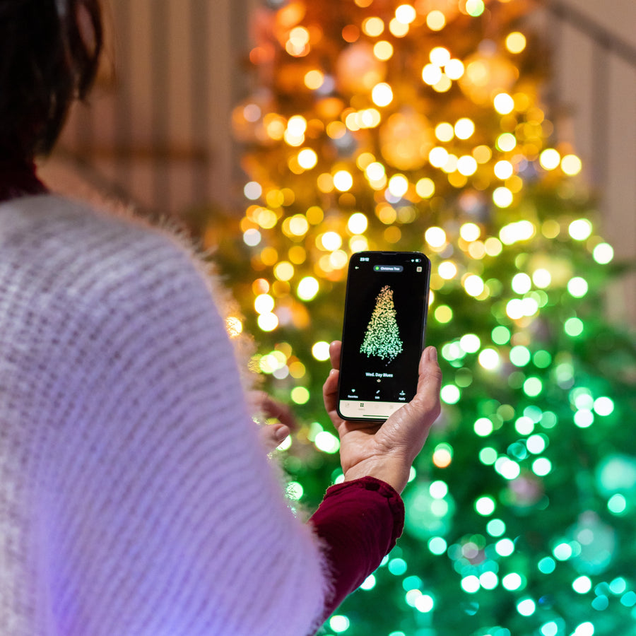 Person in weißem Pullover hält ein Smartphone, das eine beleuchtete Weihnachtsbaum-Grafik vor einem Weihnachtsbaum zeigt.