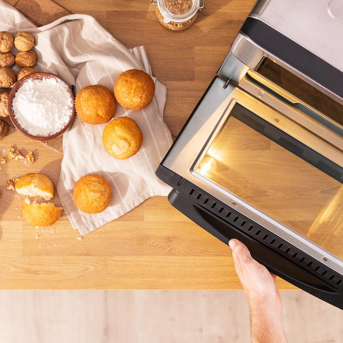 Una mano apre la porta di un forno elettrico su un piano di lavoro in legno vicino a muffin e farina.