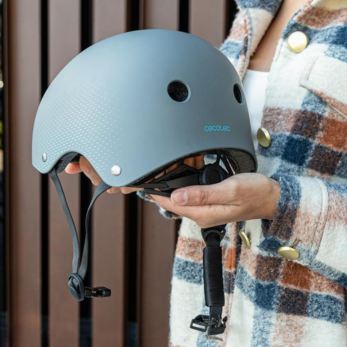 Una persona con chaqueta a cuadros sostiene un casco gris mate con correas.