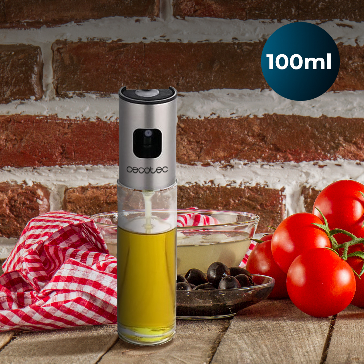 Un rociador de aceite de acero inoxidable de 100 ml sobre una mesa de madera con tomates, aceitunas y un paño a cuadros.