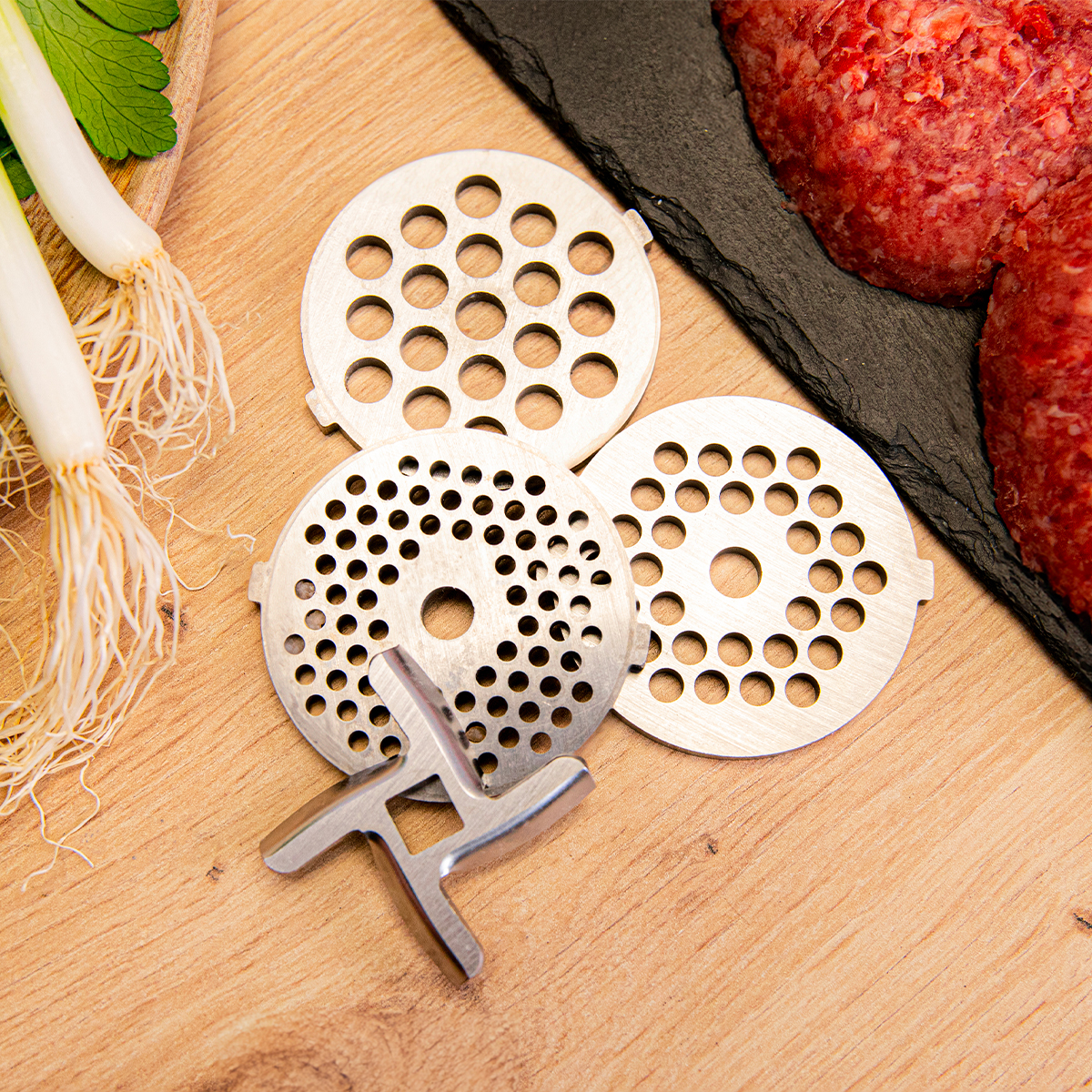 Dischi tritacarne, lama, carne macinata cruda e cipollotti su asse di legno.