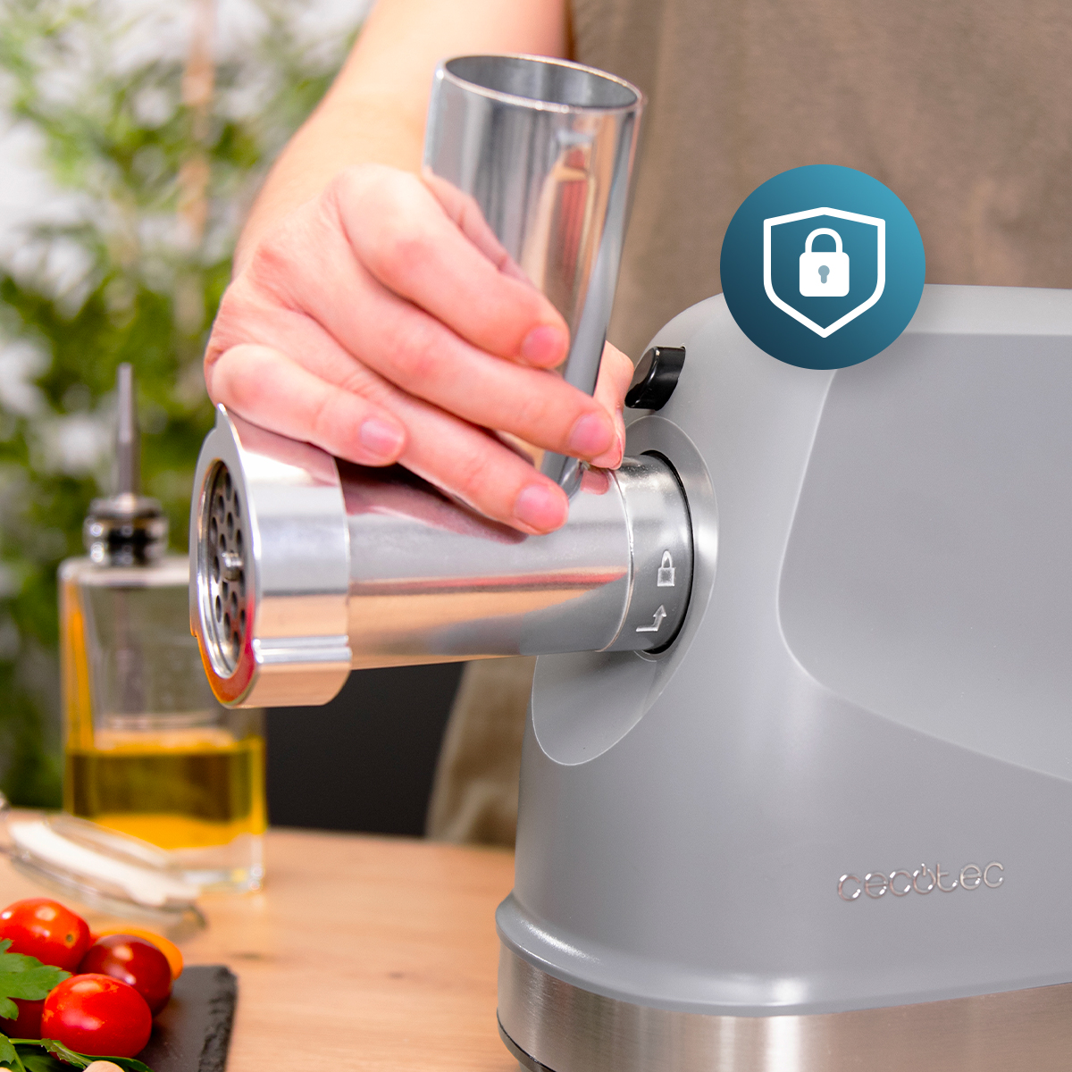Una mano fissa un accessorio a un tritacarne grigio, con olio e pomodori sullo sfondo.