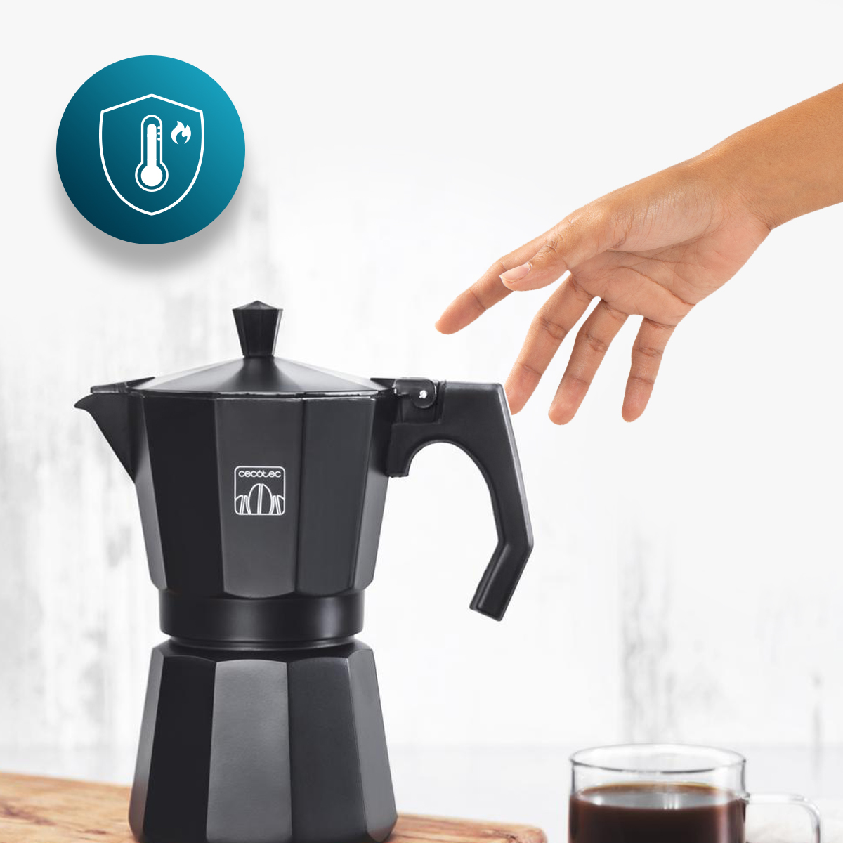 Mano acercándose a una cafetera moka negra junto a una taza de café y un icono de protección térmica.