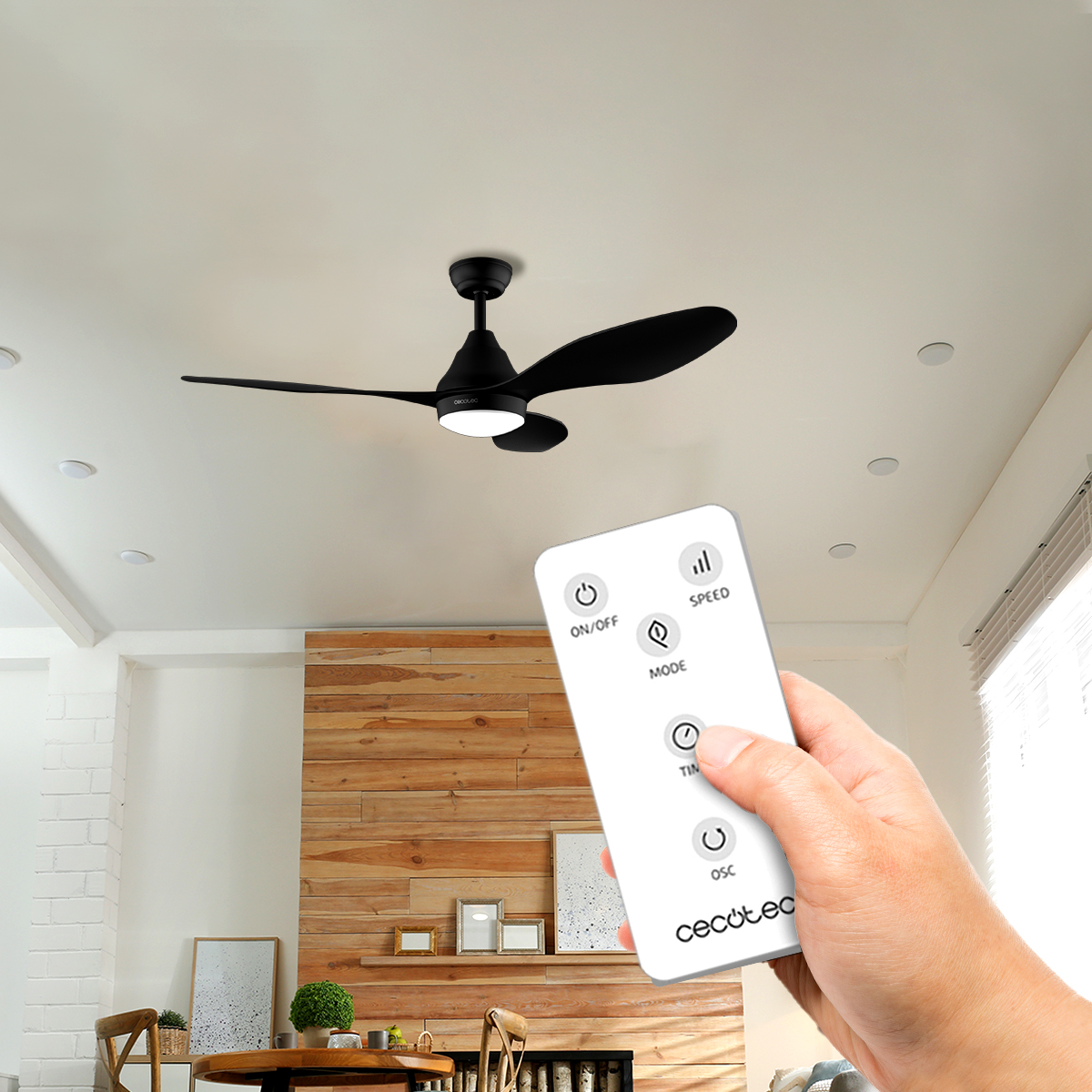 Una mano tiene un telecomando bianco in una stanza moderna, con un ventilatore da soffitto nero.