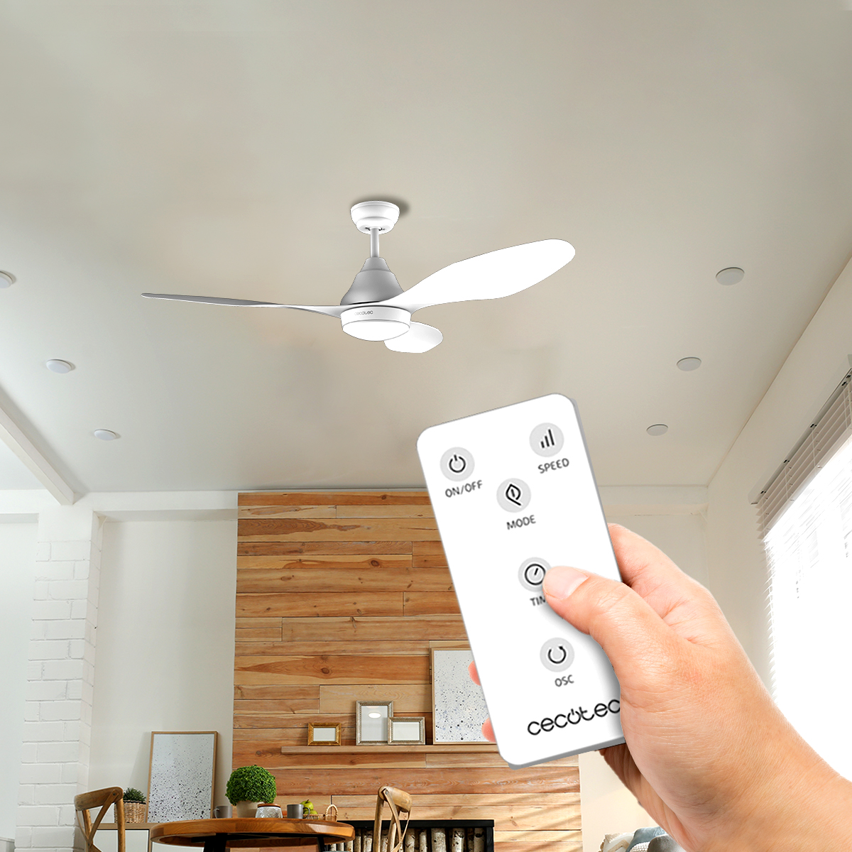 Una mano tiene un telecomando bianco davanti a un ventilatore a soffitto in un soggiorno.