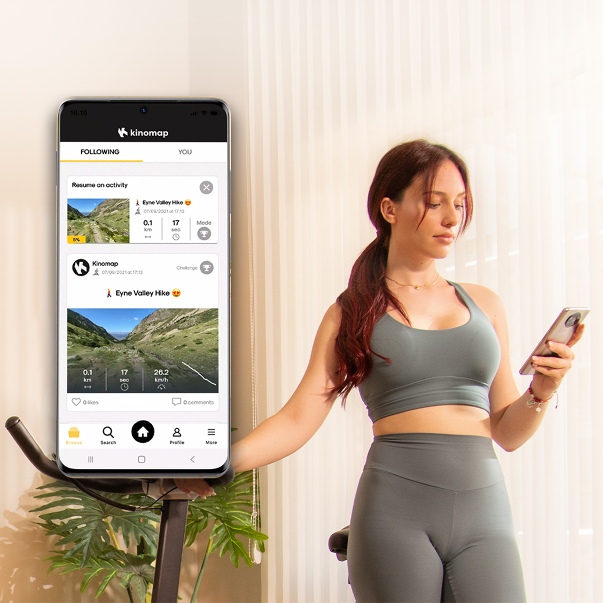 Una mujer en ropa deportiva junto a una bicicleta estática usa su teléfono con la aplicación Kinomap.