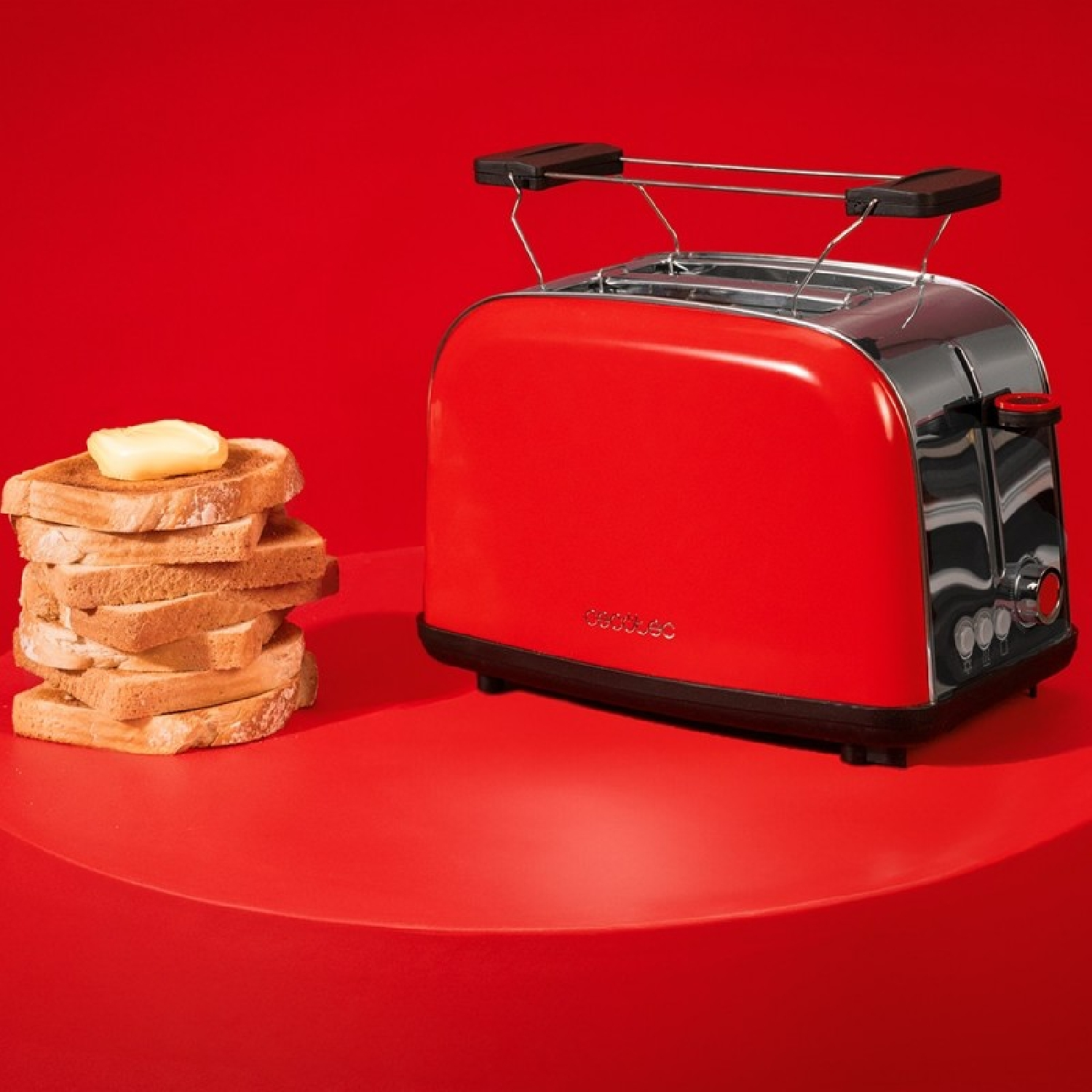 Una tostadora roja sobre una mesa roja junto a una pila de pan tostado con mantequilla.