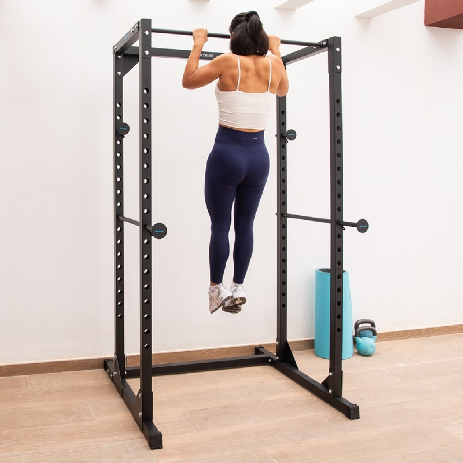 Una mujer con top blanco y mallas azules hace dominadas en una jaula de potencia negra en una habitación luminosa.