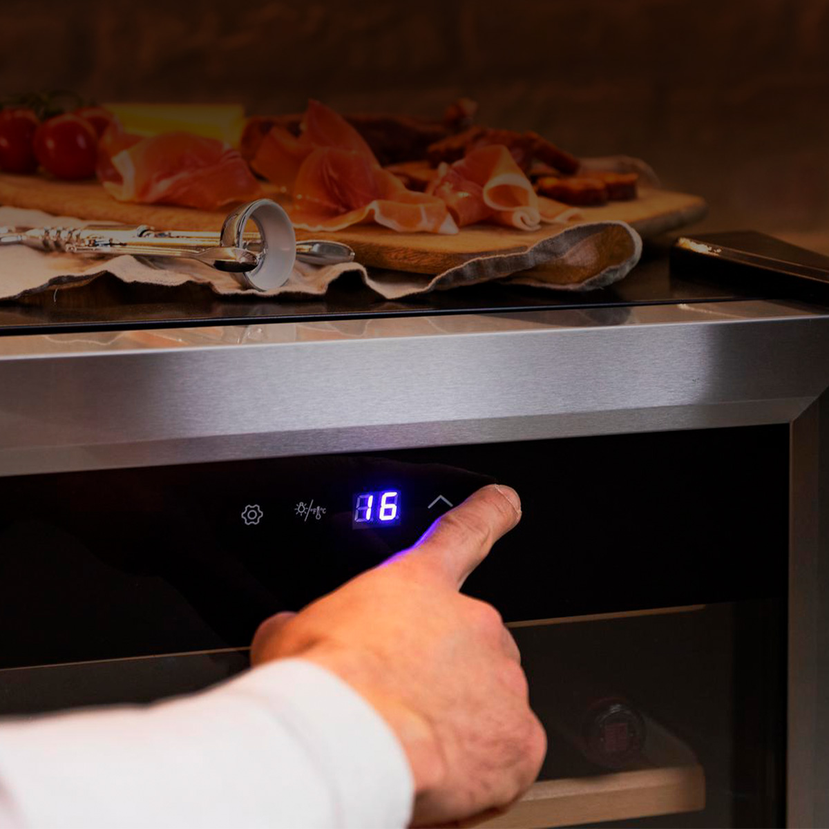 Una mano tocca il display LED blu di una cantinetta per vino, con del cibo su un asse di legno sullo sfondo.