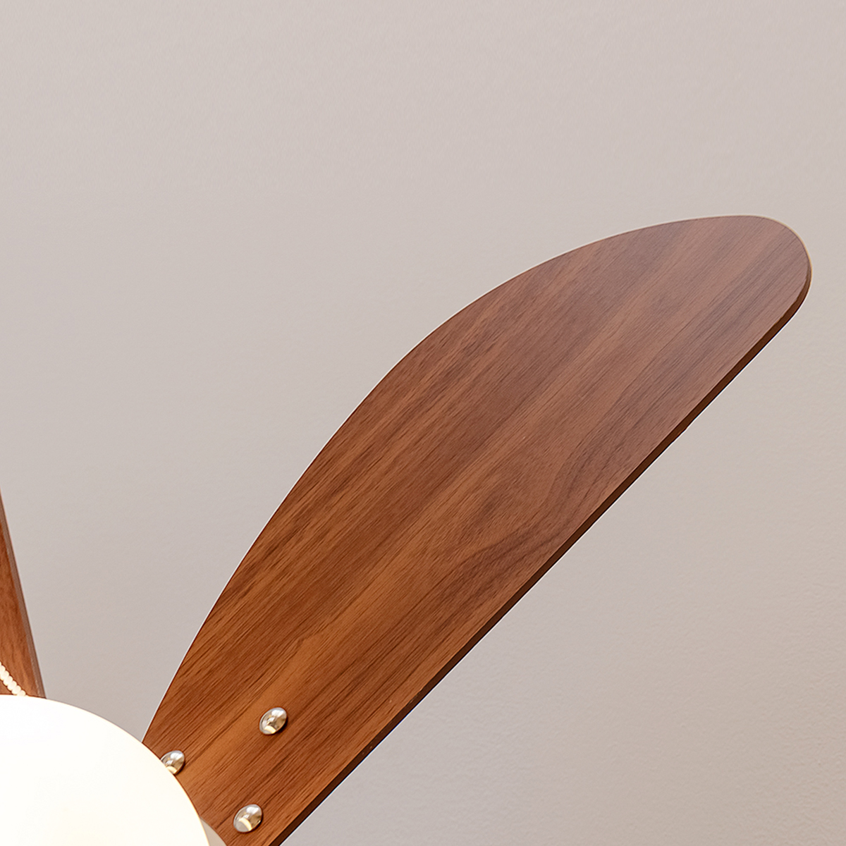 Nahaufnahme eines Holzflügels eines Deckenventilators an einer beleuchteten Leuchte vor einer hellen Wand.