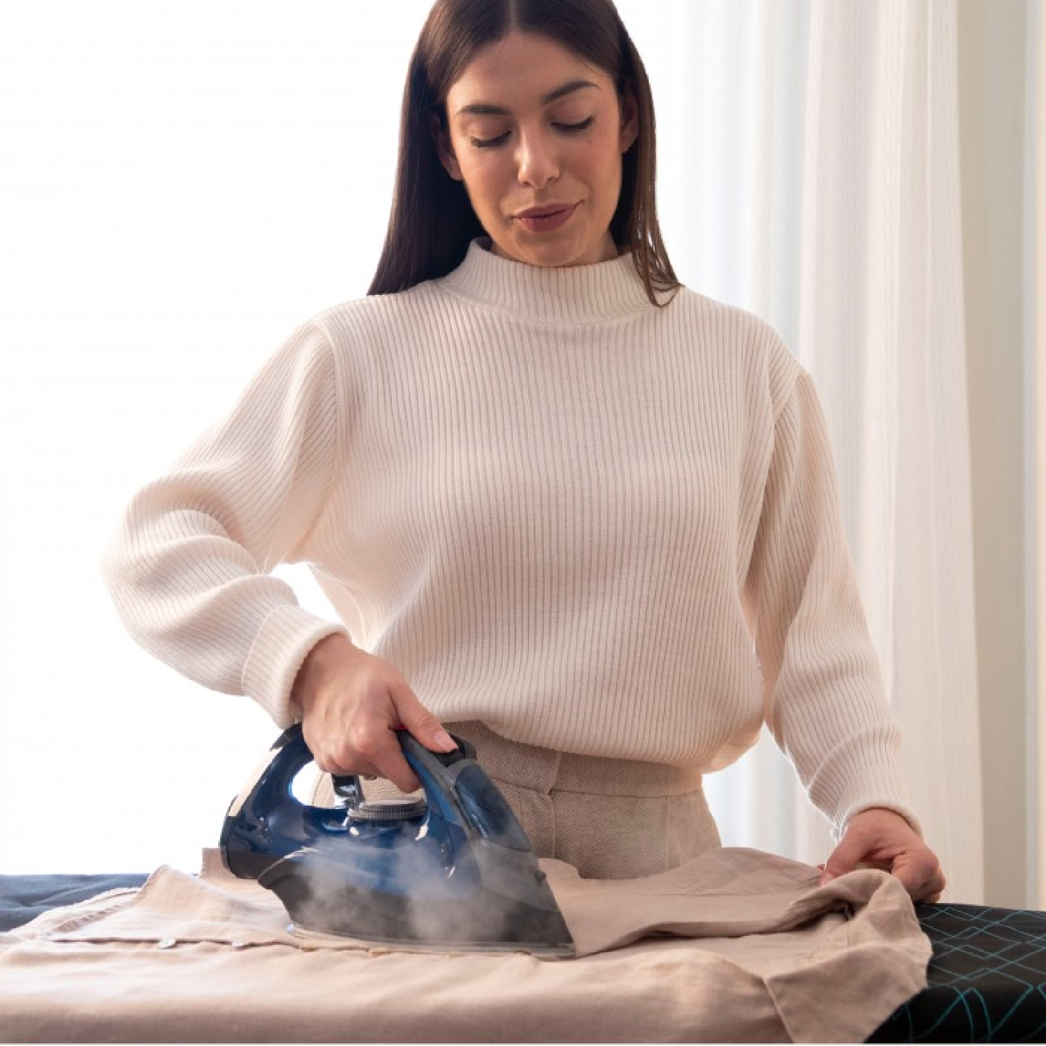 Una donna con un maglione bianco stira i vestiti su un asse da stiro con un ferro da stiro a vapore blu.