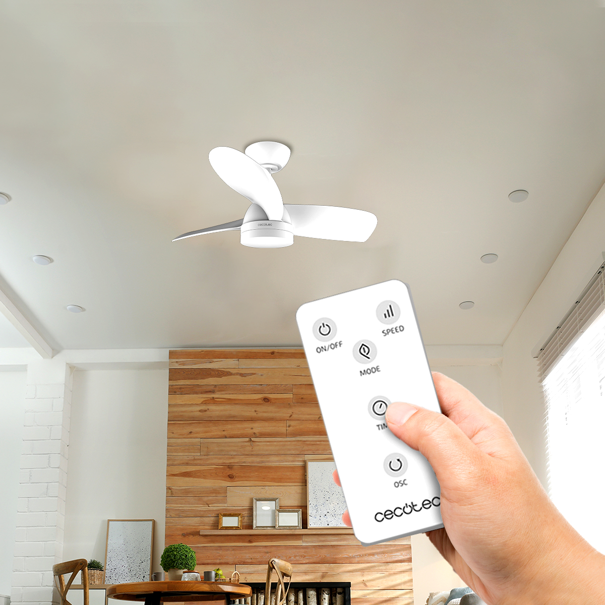 Una mano tiene un telecomando bianco per un moderno ventilatore a soffitto in una stanza con parete in legno.
