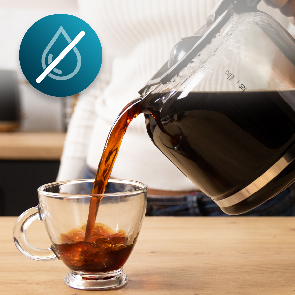 Una persona vierte café oscuro de una jarra de vidrio en una taza transparente sobre una mesa de madera con icono de gota.