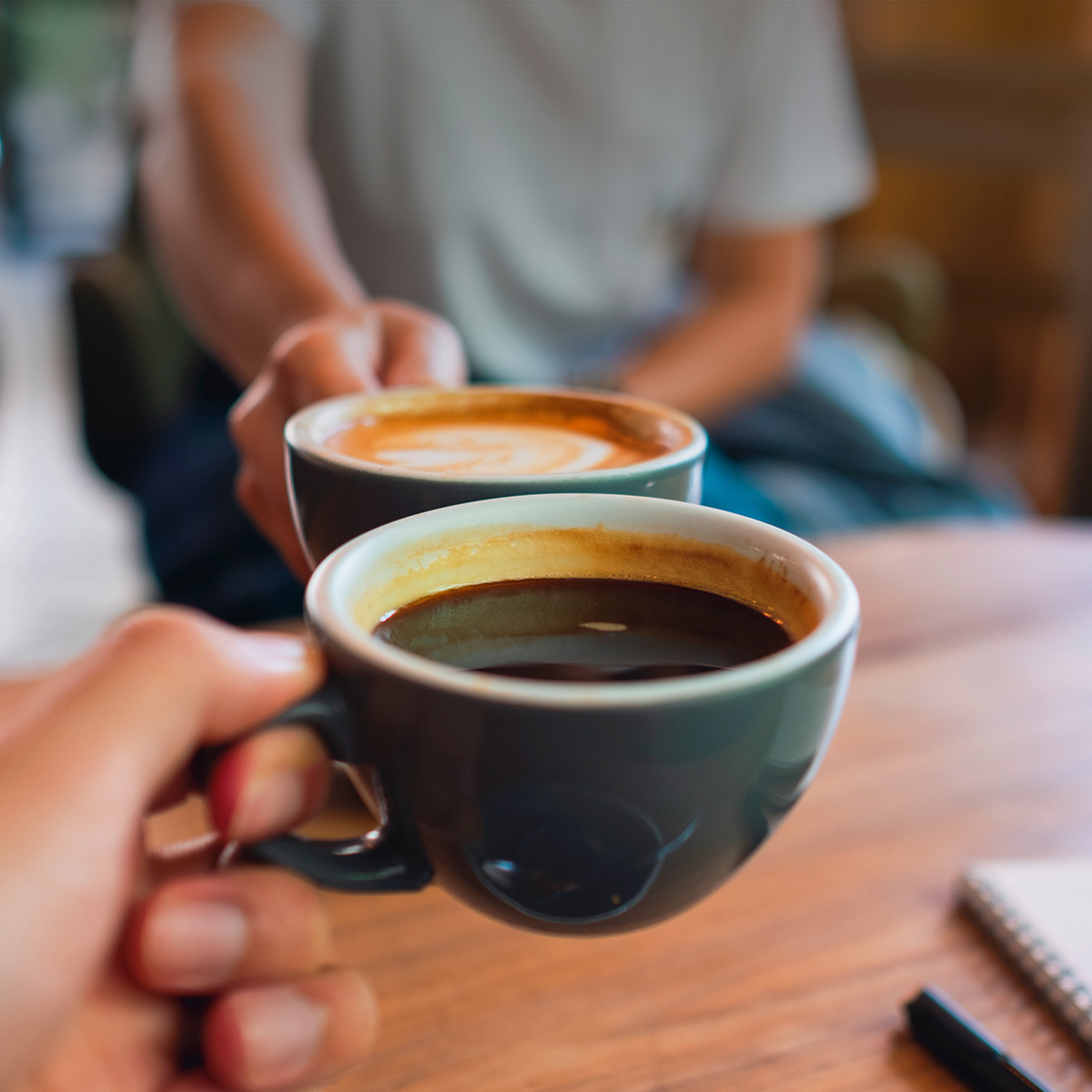 Dos personas brindando con tazas de café en una mesa de madera.