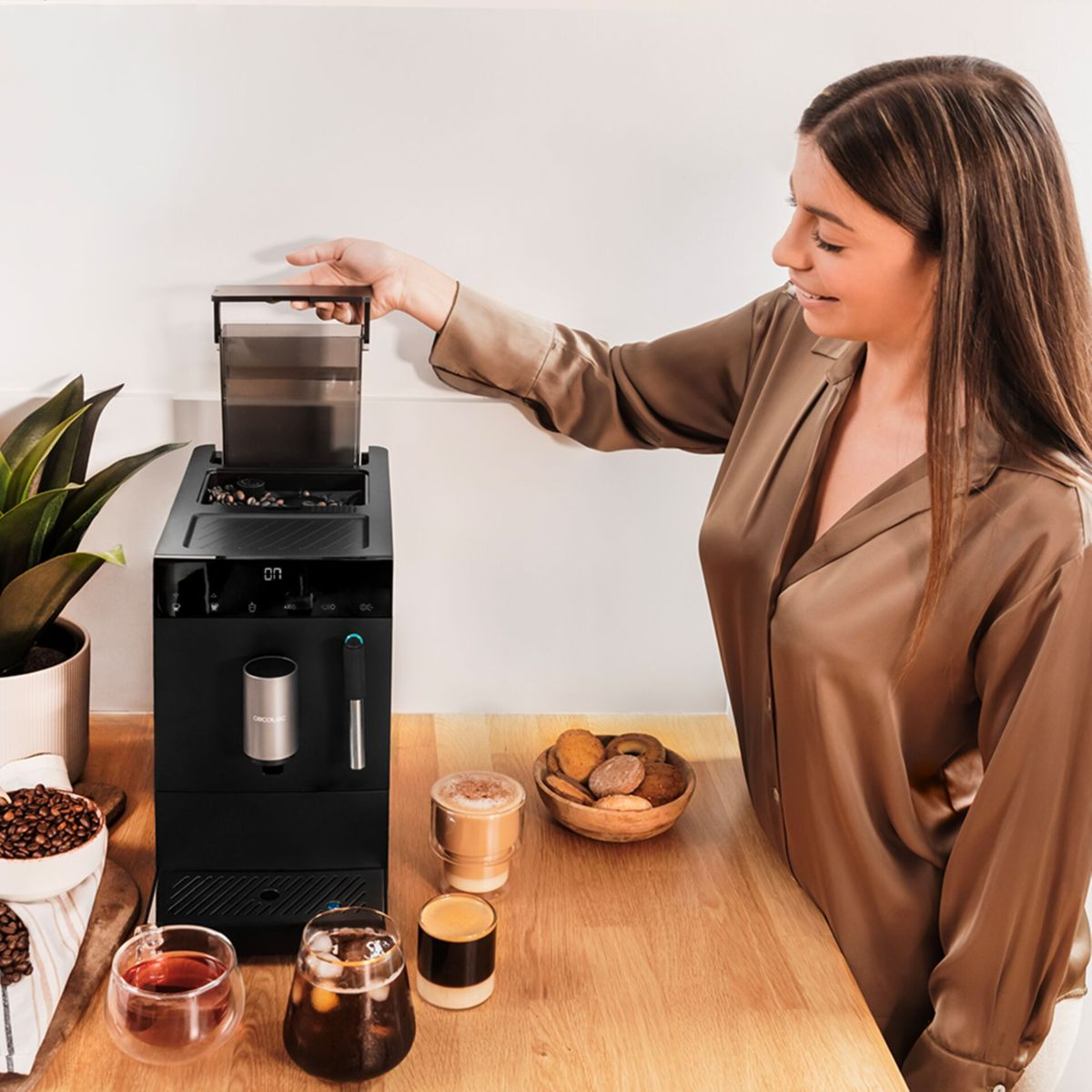 Una mujer abre una cafetera negra en una barra de madera junto a bebidas, galletas y una planta.