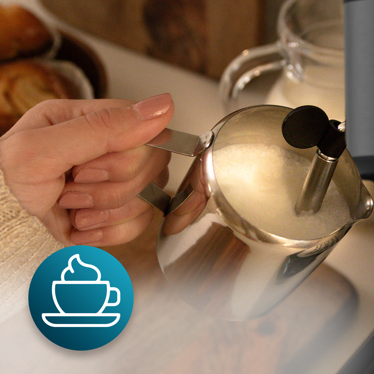 Una mano sostiene una jarra de leche de acero inoxidable con leche, hay un icono de café abajo a la izquierda.