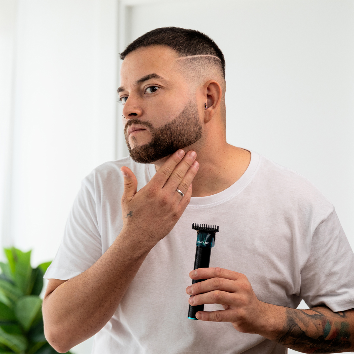 Un uomo con la barba e una maglietta bianca tiene un rifinitore e si tocca il viso.